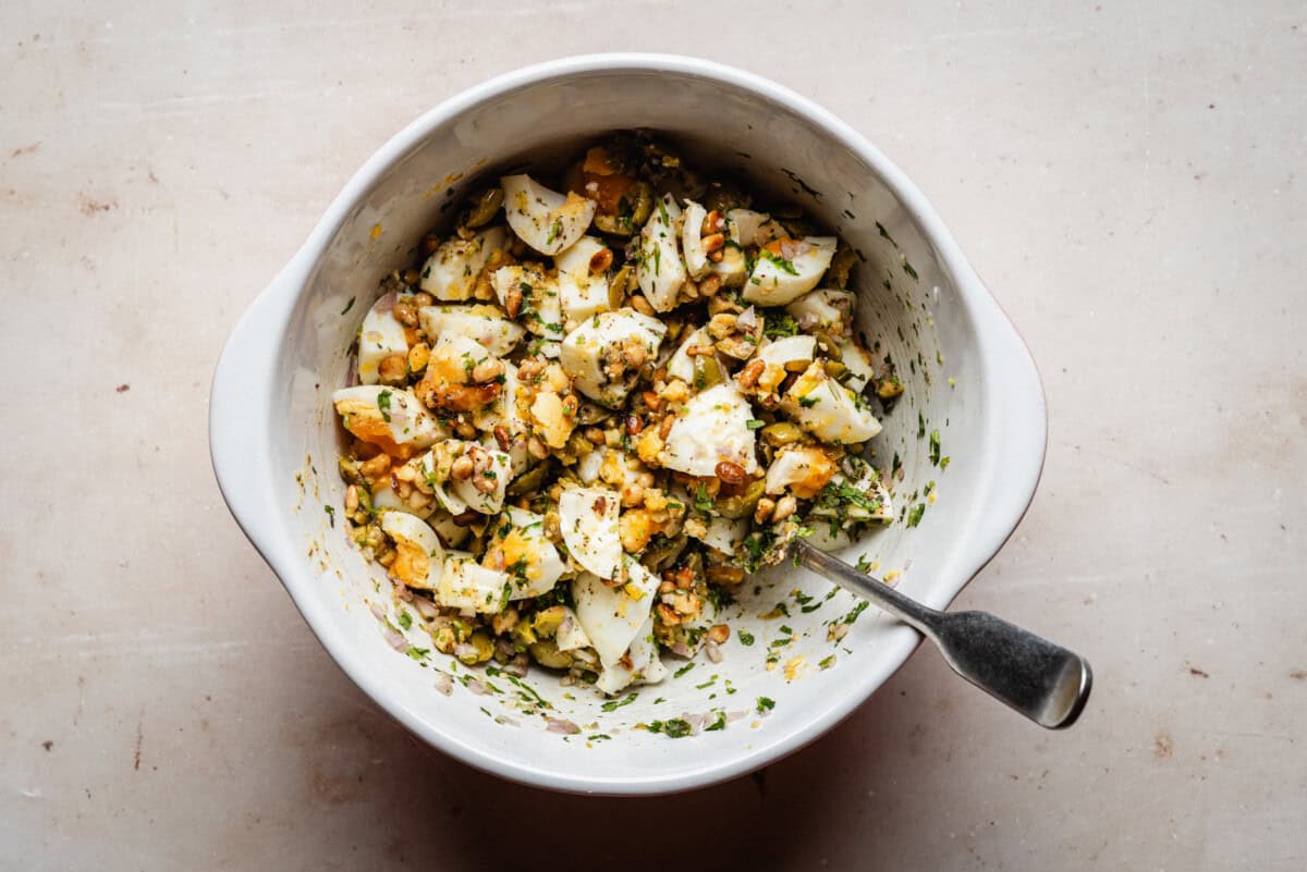 A white bowl filled with chopped hard-boiled eggs mixed with herbs and seasonings, with a spoon resting inside. The bowl sits on a light-colored surface.