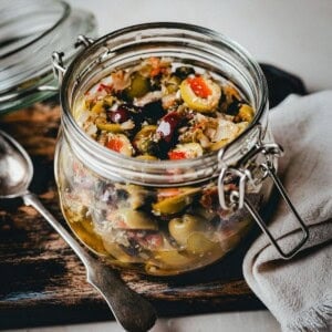 A glass jar filled with a colorful mix of chopped olives, red peppers, onions, and herbs sits on a wooden board next to a napkin and a spoon. The jar's lid is open, showcasing the vibrant medley inside.