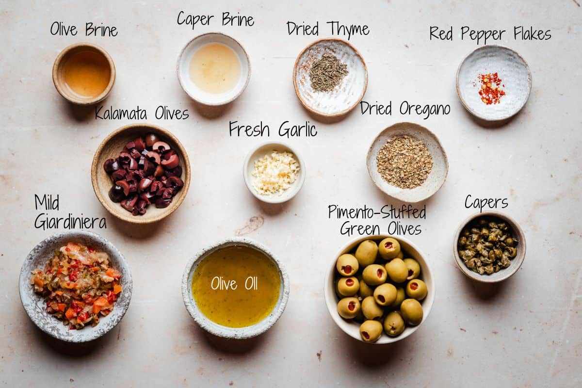 A flat lay of bowls containing olive brine, caper brine, dried thyme, red pepper flakes, kalamata olives, fresh garlic, dried oregano, mild giardiniera, olive oil, pimento-stuffed green olives, and capers, each labeled.