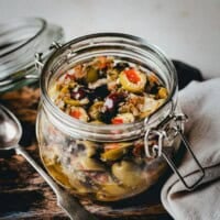 A glass jar filled with muffaletta olive salad sits on a wooden board next to a spoon and a folded napkin. The jar is open and filled with a colorful mix of chopped olives and vegetables.