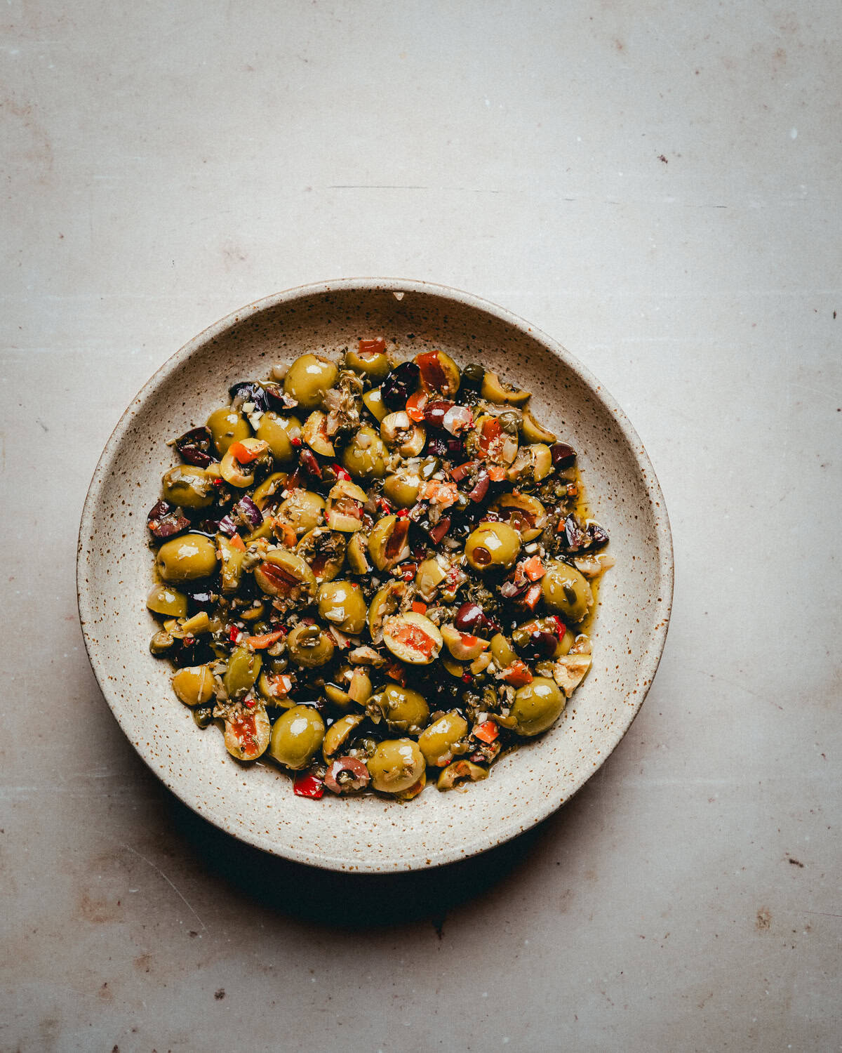A beige bowl filled with Muffaletta olive salad, including green and black olives, red chili, and herbs, placed on a light-colored textured surface.