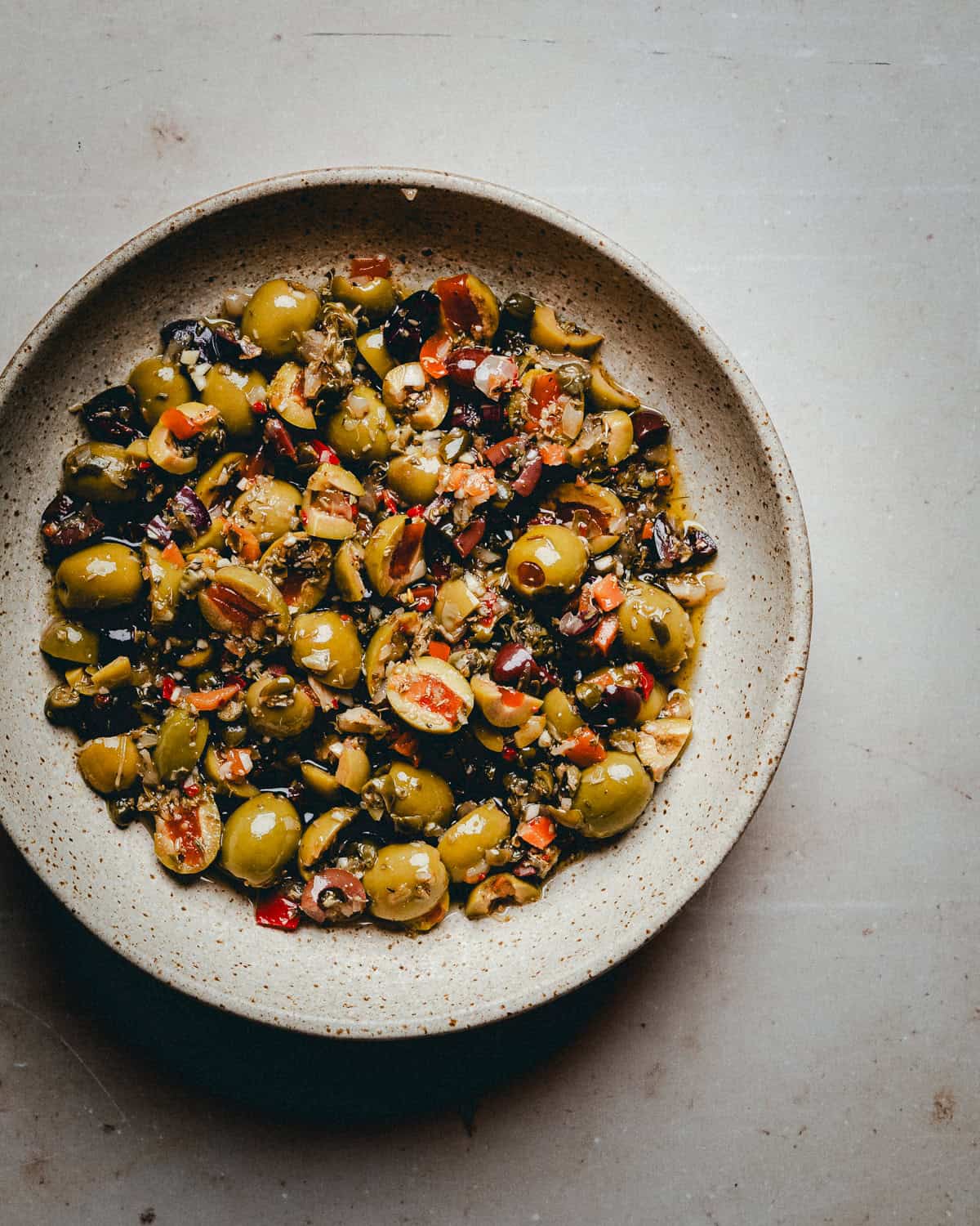 A ceramic bowl filled with Muffaletta olive salad, featuring green and black olives, diced red peppers, and herbs, sits on a light gray surface.