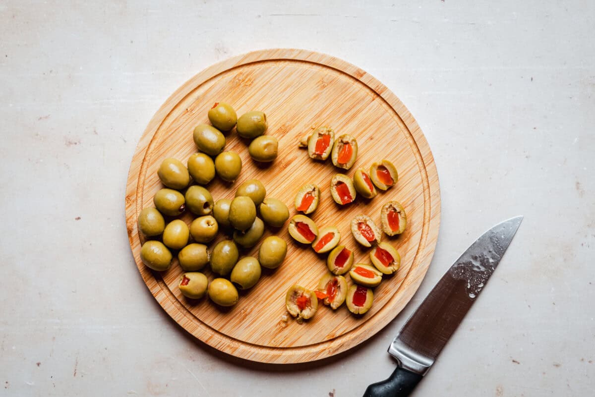 A round wooden cutting board with whole green olives on one side and sliced, pimento-stuffed green olives on the other, next to a kitchen knife on a light countertop.
