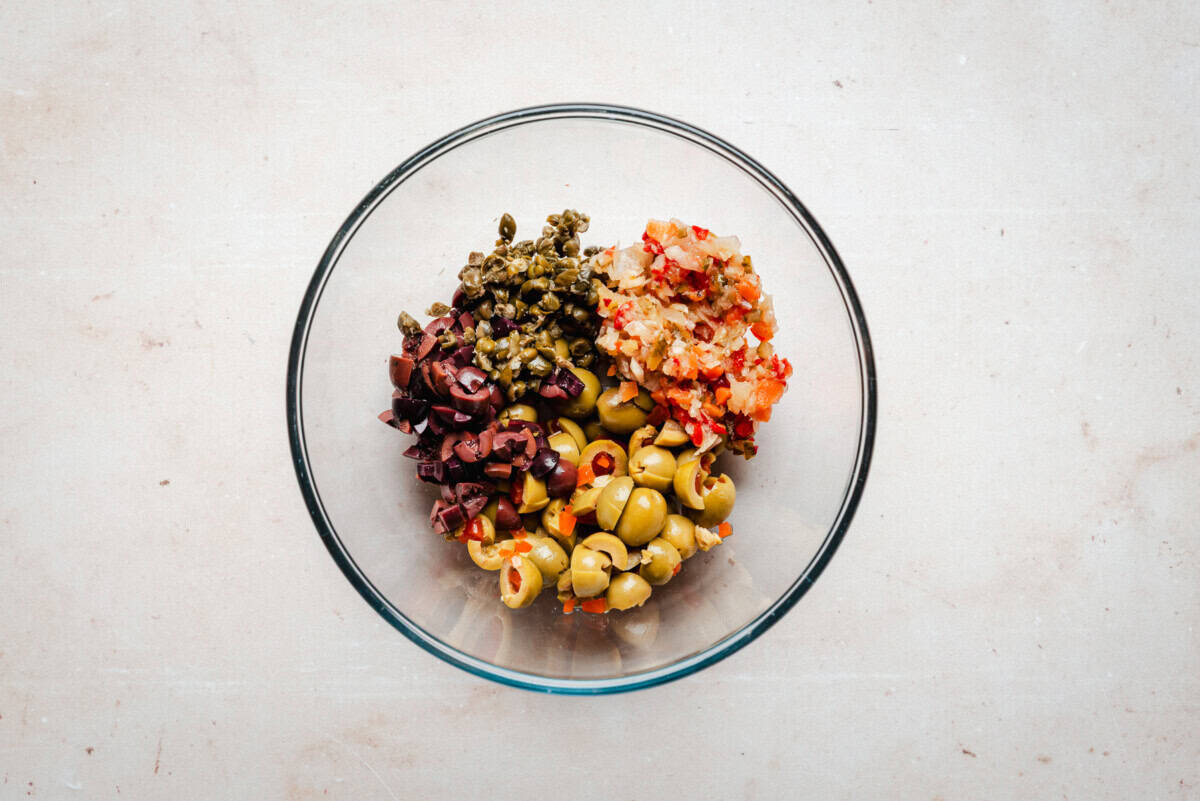 A clear glass bowl containing chopped black olives, green olives, capers, and diced red peppers, arranged in separate sections on a light-colored surface.