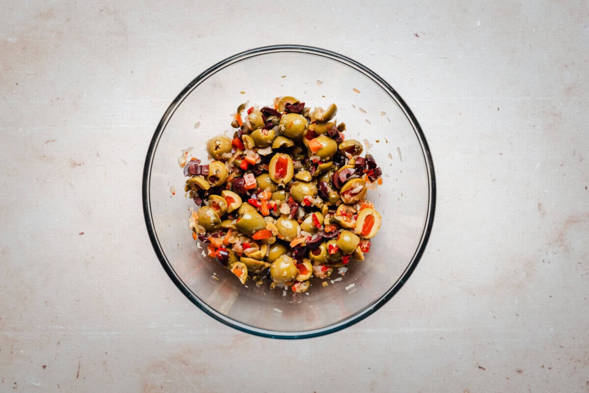 A clear glass bowl filled with a chopped olive salad mixture containing green and black olives, red peppers, and diced onions, set on a light-colored countertop.