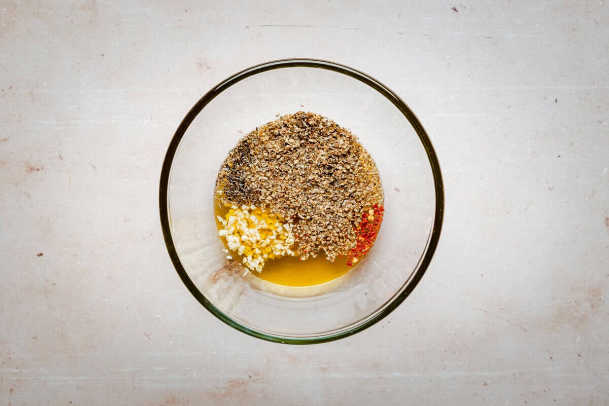 A glass bowl containing minced garlic, dried herbs, ground black pepper, red pepper flakes, and olive oil on a light-colored surface.