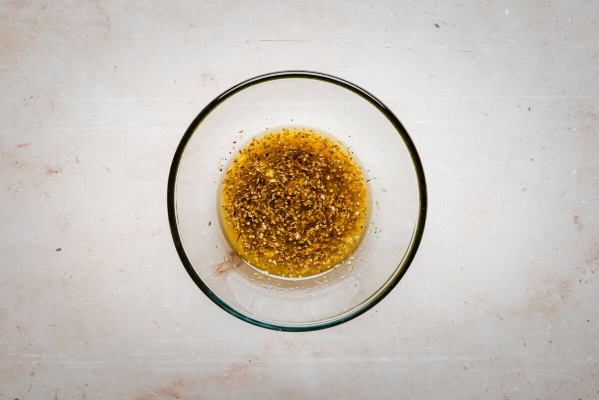 A glass bowl filled with a marinade of oil and herbs sits on a light-colored, textured surface.
