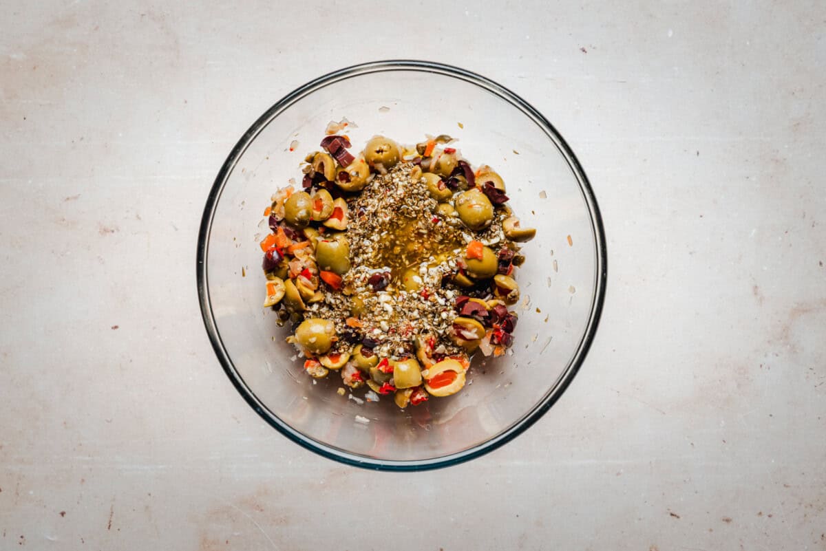 A glass bowl filled with chopped green and black olives, diced red peppers, dried herbs, and olive oil, placed on a light-colored surface.