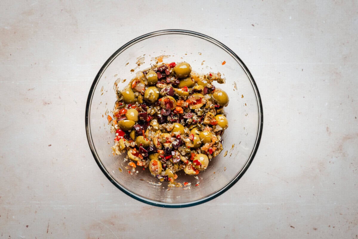 A glass bowl filled with a mixture of green and black chopped olives, diced red peppers, and seasonings on a light, neutral surface.