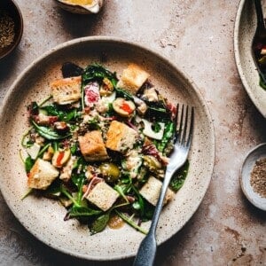 A plate of fresh Muffaletta Salad with leafy greens, tomatoes, olives, and croutons, topped with dressing. A fork rests on the plate, and there are bowls of dressing and seeds nearby on a rustic surface.