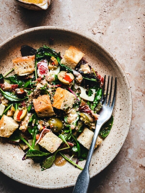 A plate of fresh Muffaletta Salad with leafy greens, tomatoes, olives, and croutons, topped with dressing. A fork rests on the plate, and there are bowls of dressing and seeds nearby on a rustic surface.