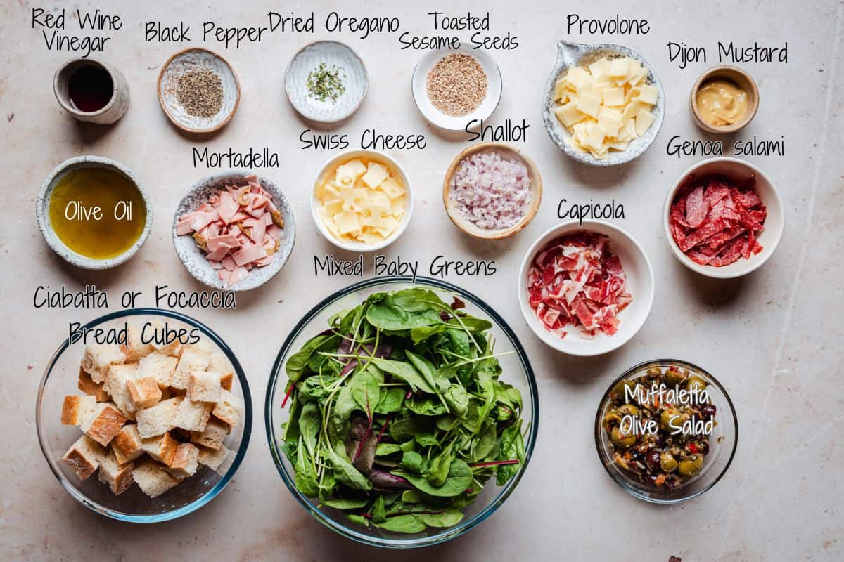 A flat lay of ingredients for Muffaletta Salad, including bowls of mixed greens, cubed bread, various meats, cheeses, shallot, seasonings, olive oil, vinegar, Dijon mustard, and muffuletta olive salad, all labeled.
