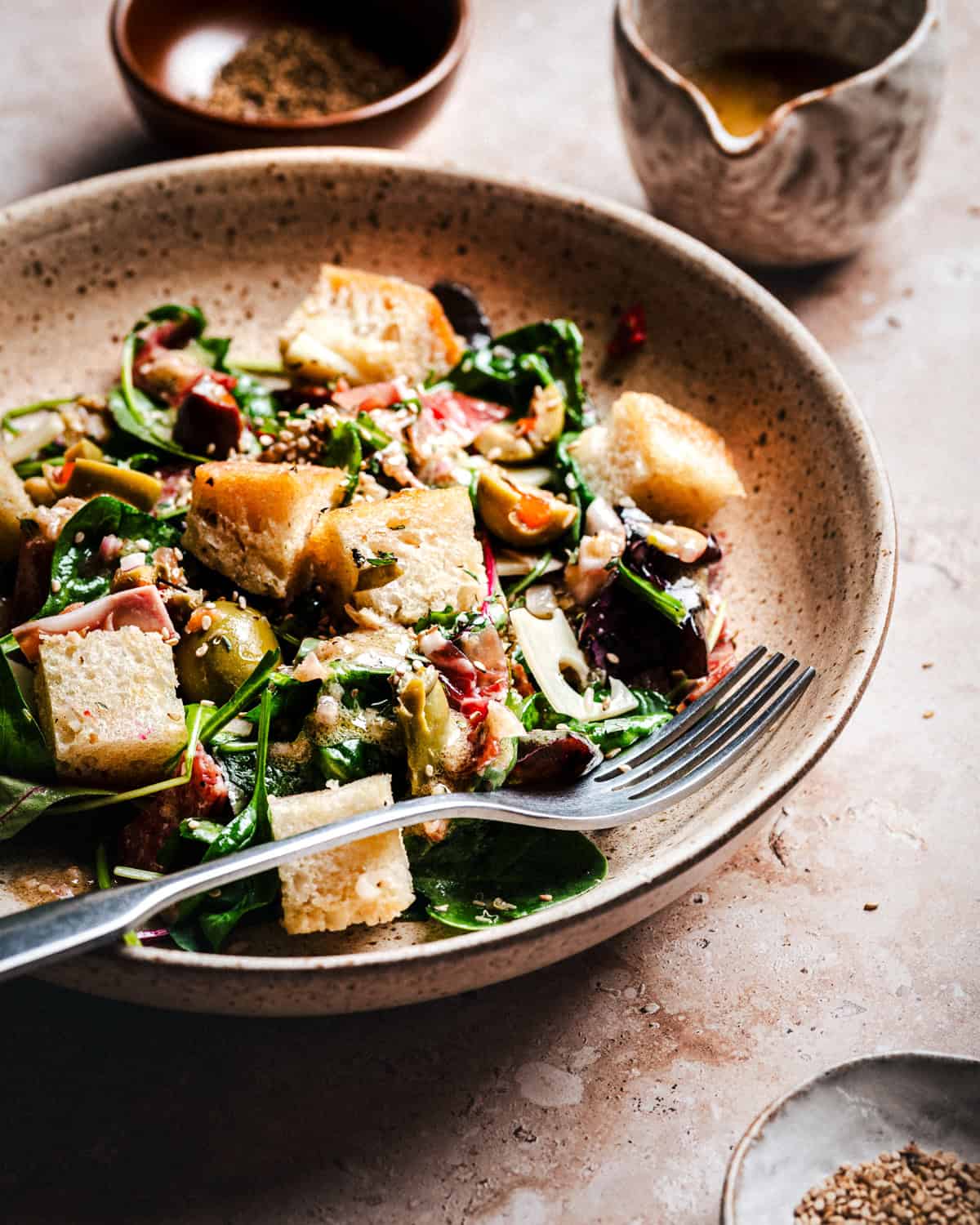 A bowl of bread fresh salad with green, crouton, and olives, served with a fork. In the background, there are small bowls containing dressing and spices.