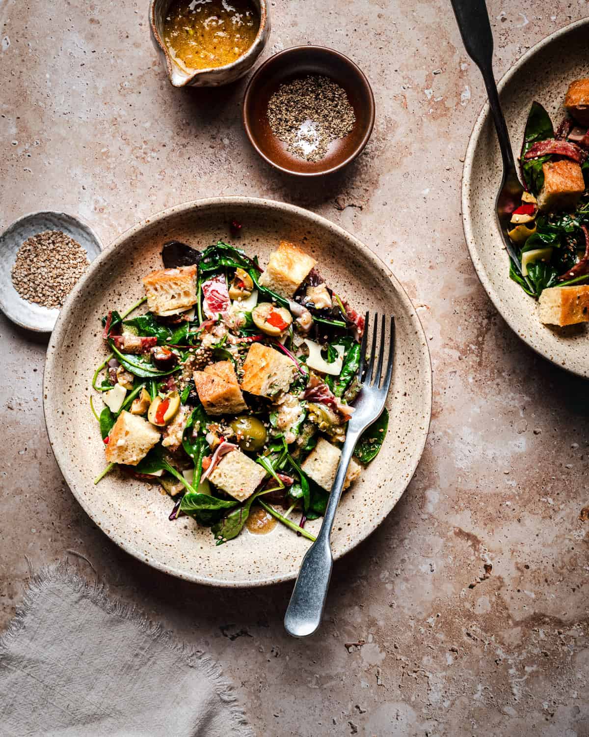 A plate of bread salad with leafy greens, olives, and cheese, served with a fork. Small bowls of dressing and pepper are nearby on a light stone surface.