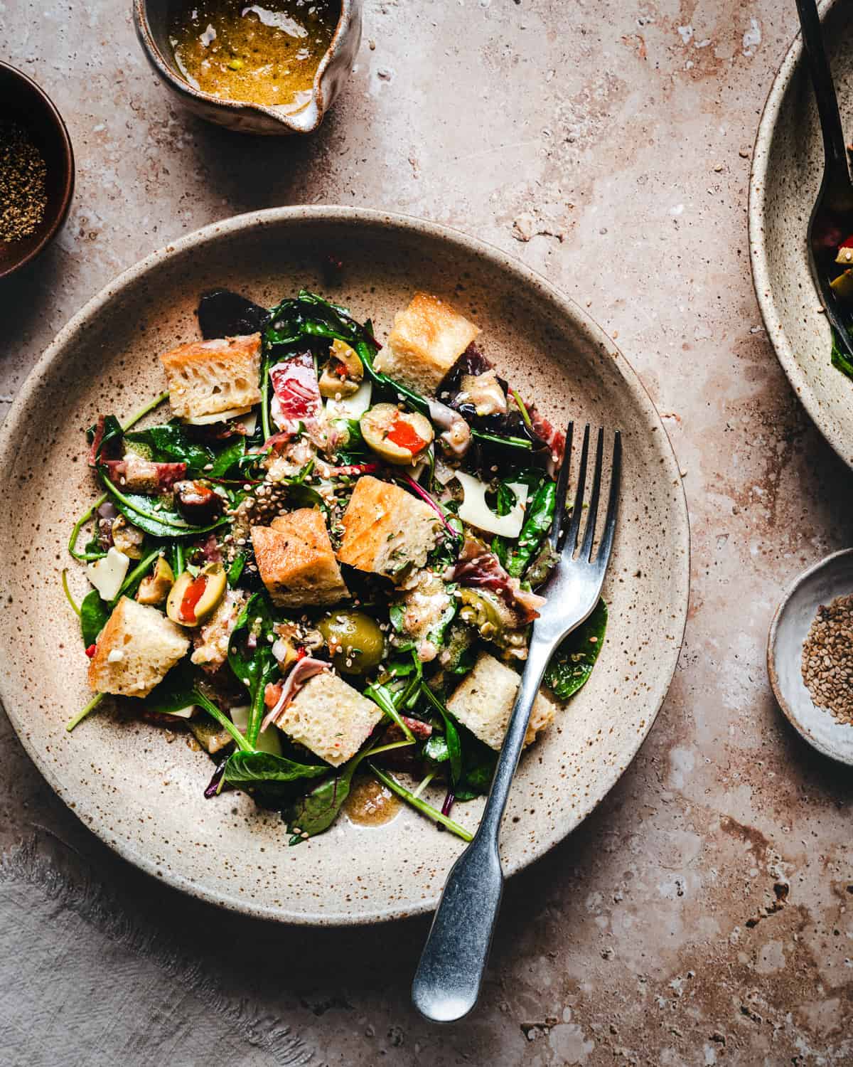 A plate of fresh bread salad with mixed greens, olives, and sesame seeds, topped with a light dressing. A fork rests on the plate, with bowls of dressing and seasoning nearby on a textured surface.