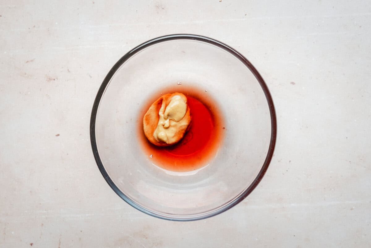 A clear glass bowl contains a dollop of mustard and red wine vinegar on a light-colored surface.
