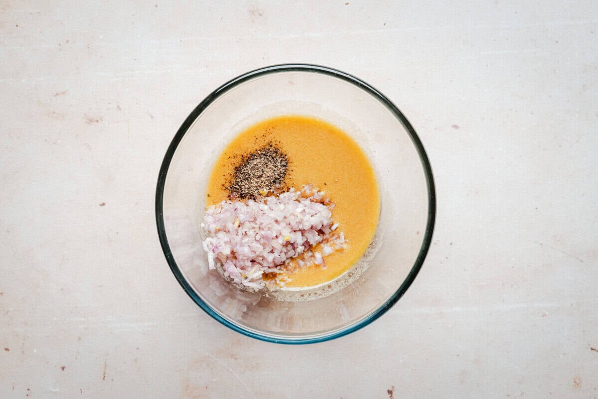 A glass bowl containing a yellow liquid, finely chopped shallots, black pepper, and salt, placed on a light-colored surface.