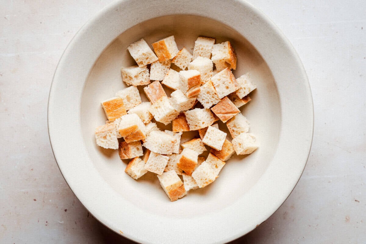 A beige bowl filled with small, evenly cut cubes of bread, placed on a light-colored surface.