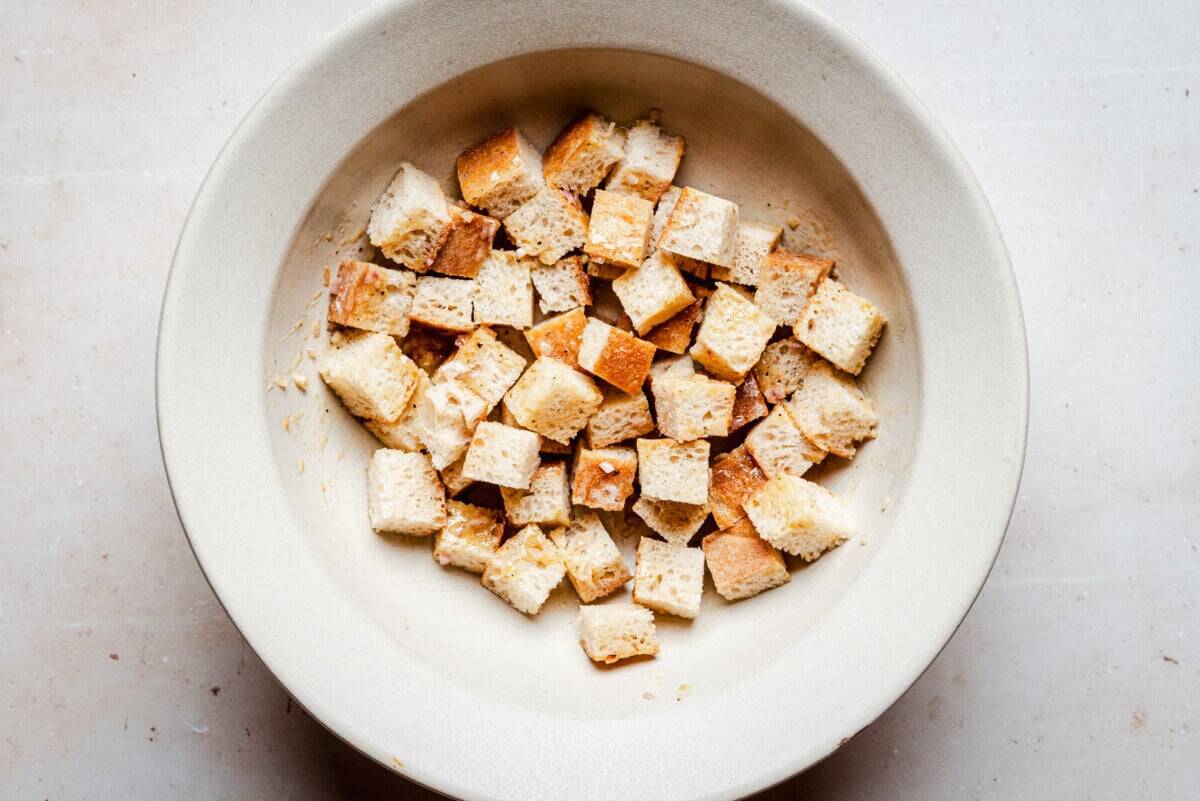 A beige bowl filled with evenly cut cubes of bread drizzled with dressing, sits on a light-colored surface.