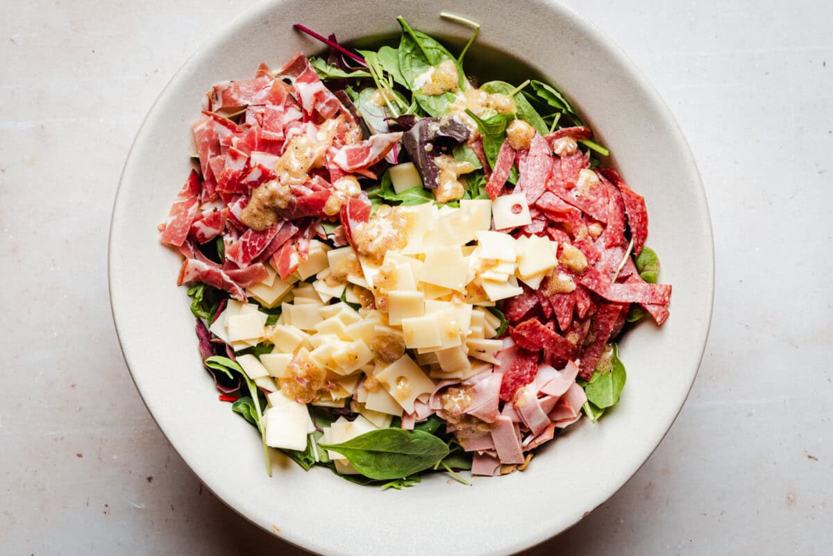 A bowl filled with mixed greens, diced cheese, salami, and prosciutto, topped with creamy dressing. The ingredients are arranged in separate sections on top of the greens.
