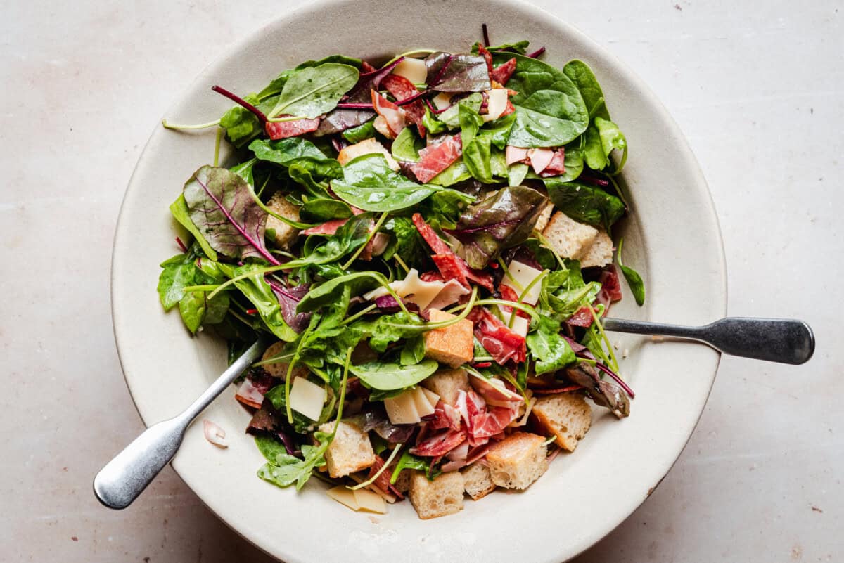 A bowl of mixed salad with leafy greens, thin cheese slices, diced cured meat, and croutons, with two metal utensils resting inside.