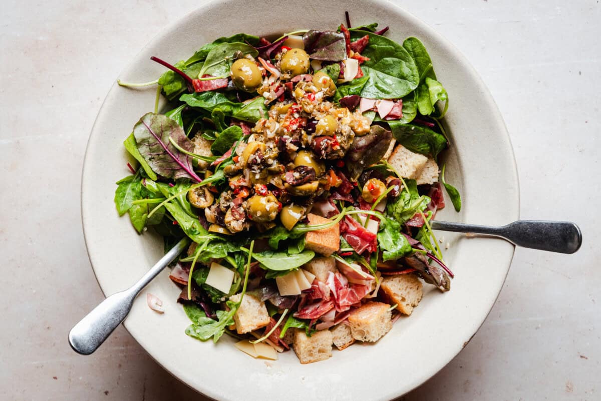 A bowl of salad with leafy greens, diced prosciutto, cheese, croutons, and a topping of chopped olives and peppers, with two serving utensils in the bowl.