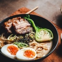 A bowl of pork belly ramen with sliced pork belly, halved soft-boiled eggs, noodles, bok choy, and seaweed. The bowl is on a brown surface with chopsticks and a pinchandswirl.com banner at the bottom.