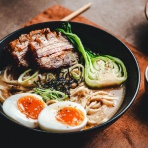 A bowl of ramen with sliced pork belly, half-boiled eggs, bok choy, noodles, green onions, and seaweed in broth, served on a brown napkin with wooden chopsticks to the side.