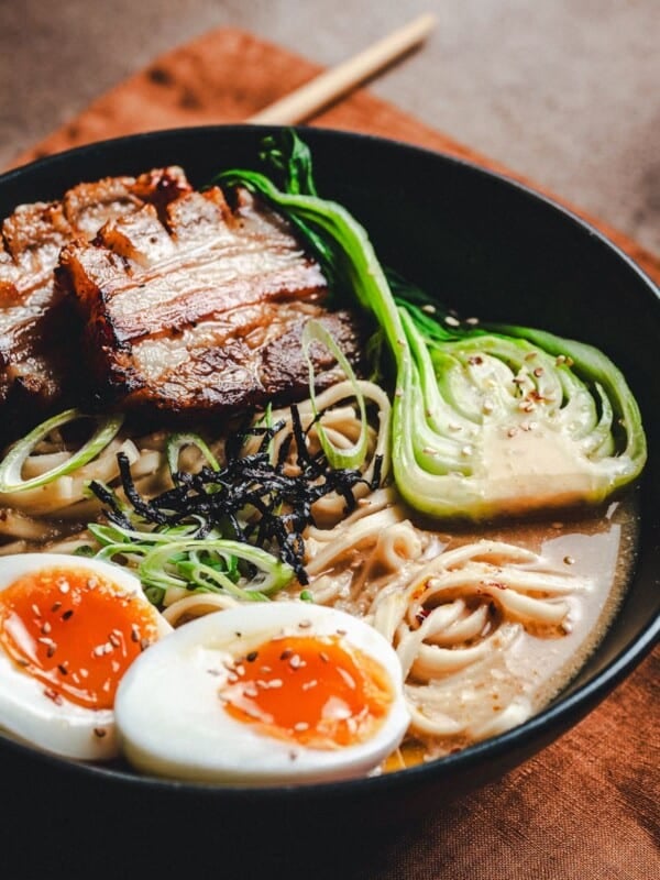 A bowl of ramen with sliced pork belly, half-boiled eggs, bok choy, noodles, green onions, and seaweed in broth, served on a brown napkin with wooden chopsticks to the side.