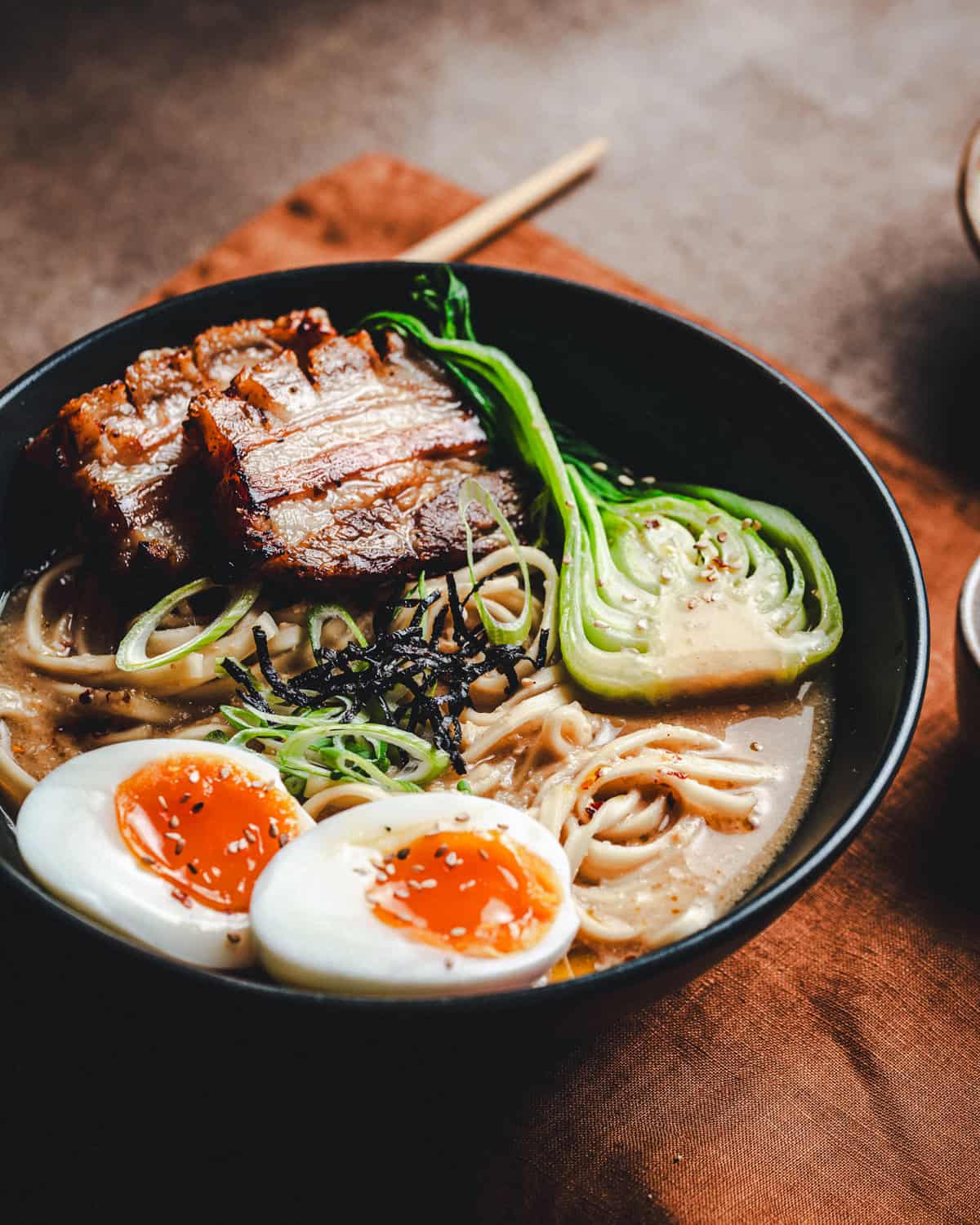 A bowl of pork belly ramen topped with two halves of soft-boiled eggs, slices of seared pork belly, bok choy, seaweed strips, and green onions, all in a rich broth with noodles. Chopsticks rest nearby on a brown cloth.