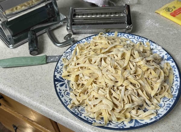 A blue and white plate filled with fresh, uncooked pasta sits on a kitchen counter next to a pasta maker, a green-handled knife, and a packet of flour.