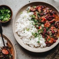 A bowl of red beans and rice with sausage, garnished with chopped green onions and parsley, sitting on a gray surface next to a small bowl of sliced green onions.