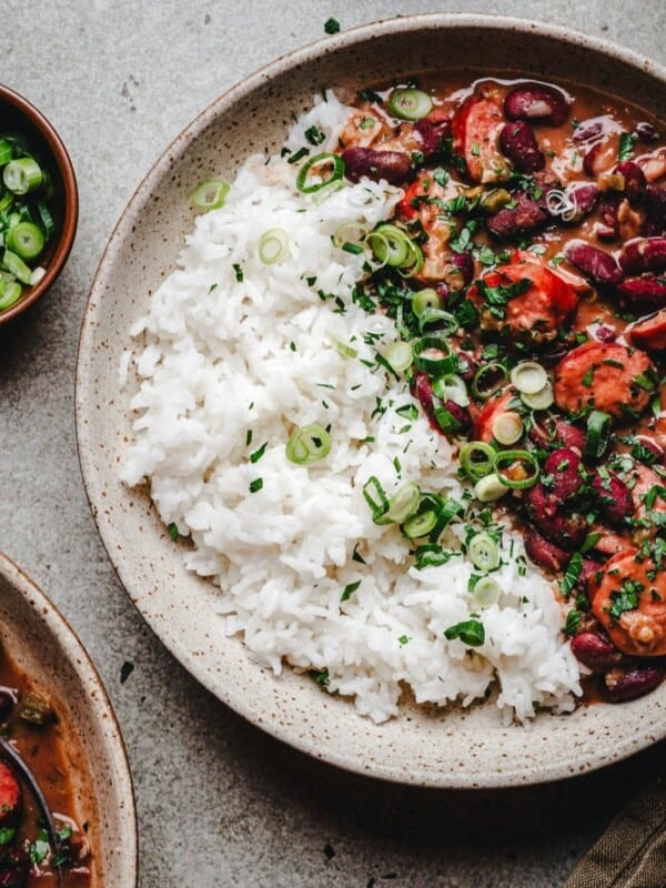 A bowl of red beans and rice with sausage, garnished with chopped green onions and parsley, sitting on a gray surface next to a small bowl of sliced green onions.
