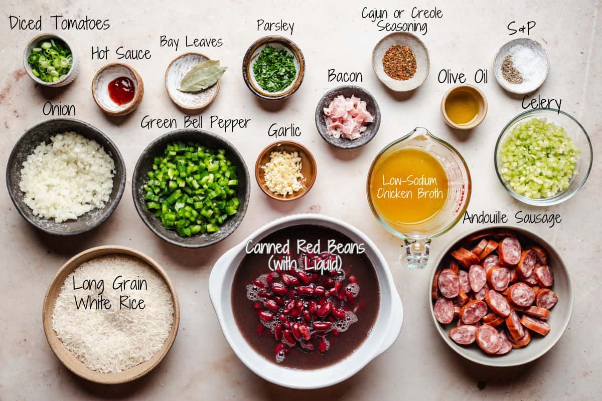 A top-down view of ingredients for red beans and rice, including diced tomatoes, onion, hot sauce, bay leaves, parsley, green bell pepper, garlic, bacon, chicken broth, sausage, celery, oil, and seasonings.