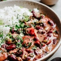 A bowl of red beans and rice topped with sliced green onions and herbs, with sausage pieces visible in the beans. The dish is served in a rustic bowl on a light surface.