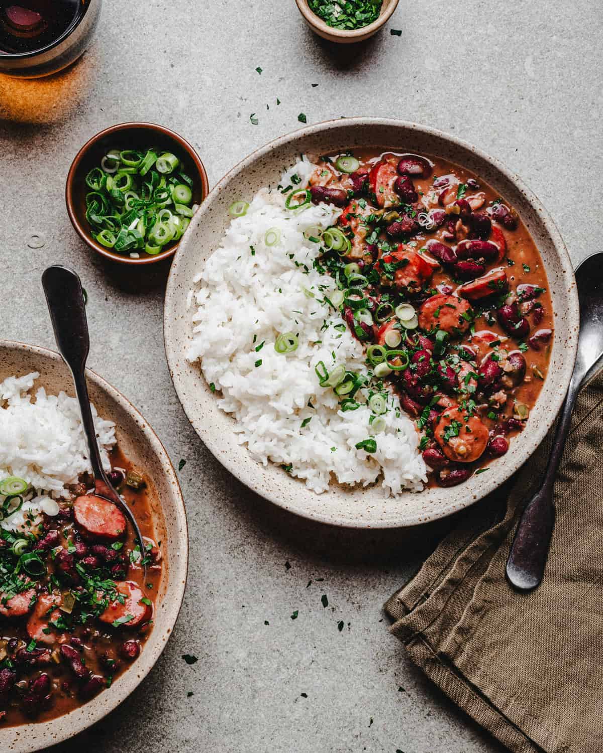 Two bowls filled with red beans and rice with sausage, and vegetables, garnished with chopped green onions and herbs, with a side of sliced green onions and a folded brown napkin nearby.