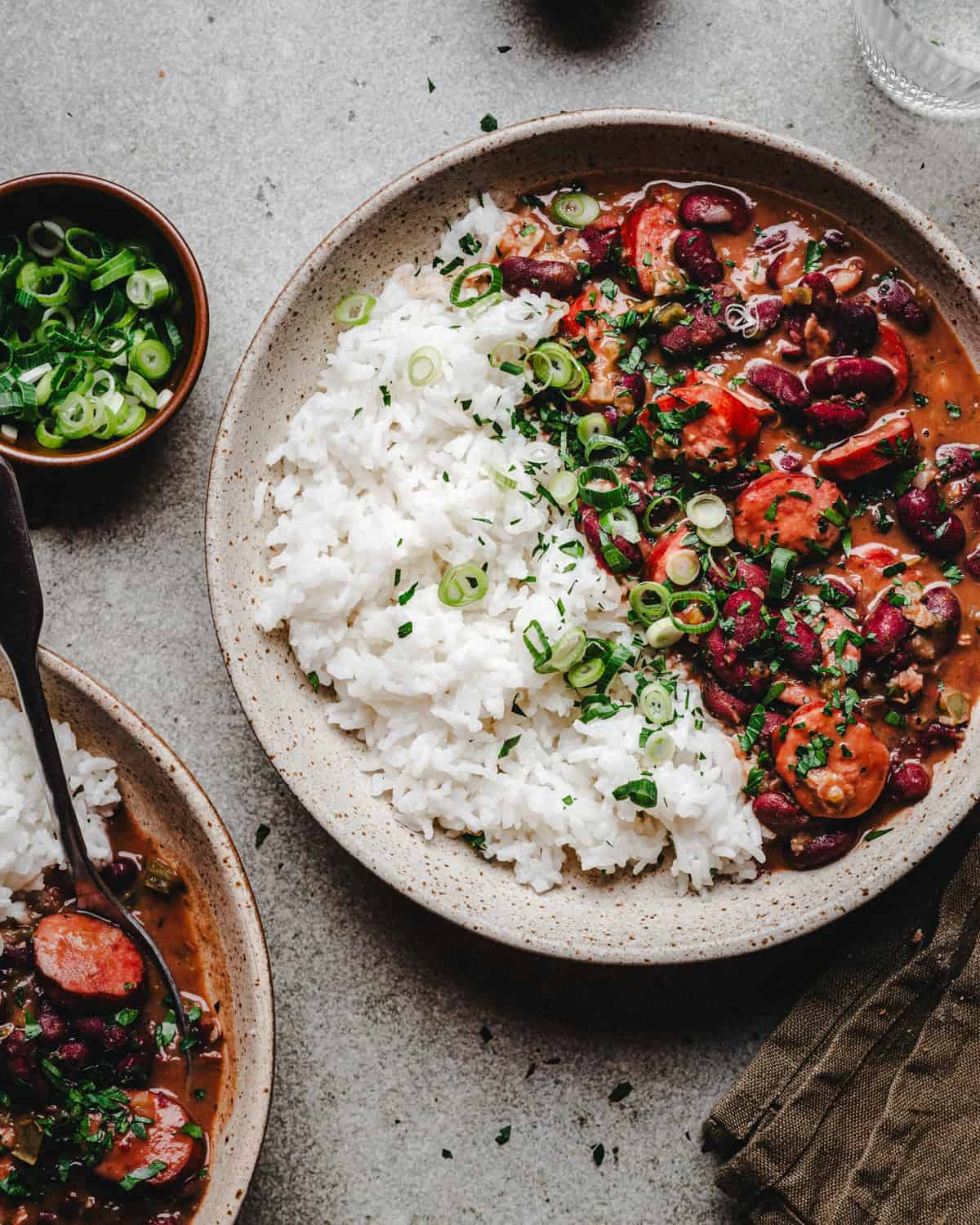 A ceramic bowl filled with red beans and rice with thick sauce, garnished with chopped green onions and herbs, sits on a gray surface next to a small bowl of sliced green onions.