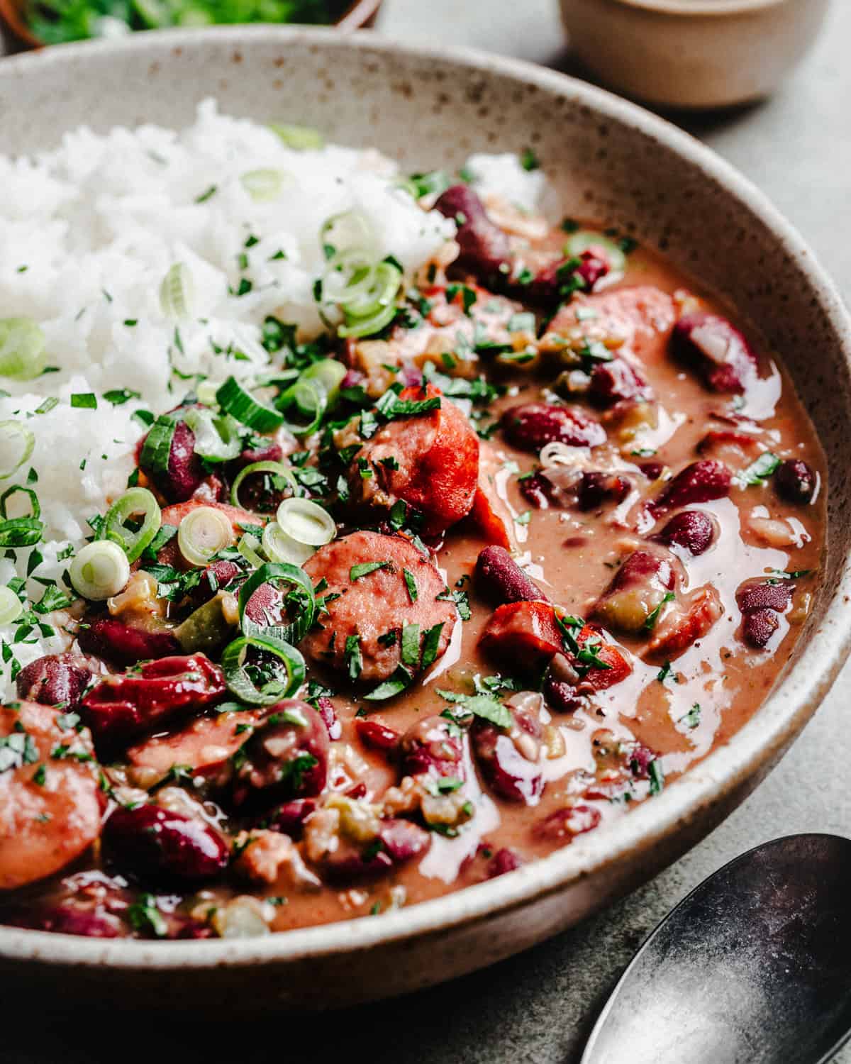 A close-up of a bowl filled with red beans and rice with sliced sausage, garnished with chopped green onions and herbs.