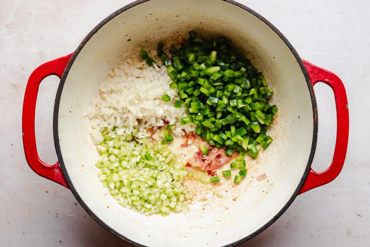 Chopped onions, celery, and green bell peppers are arranged in sections inside a red Dutch oven on a light surface, ready to be cooked.