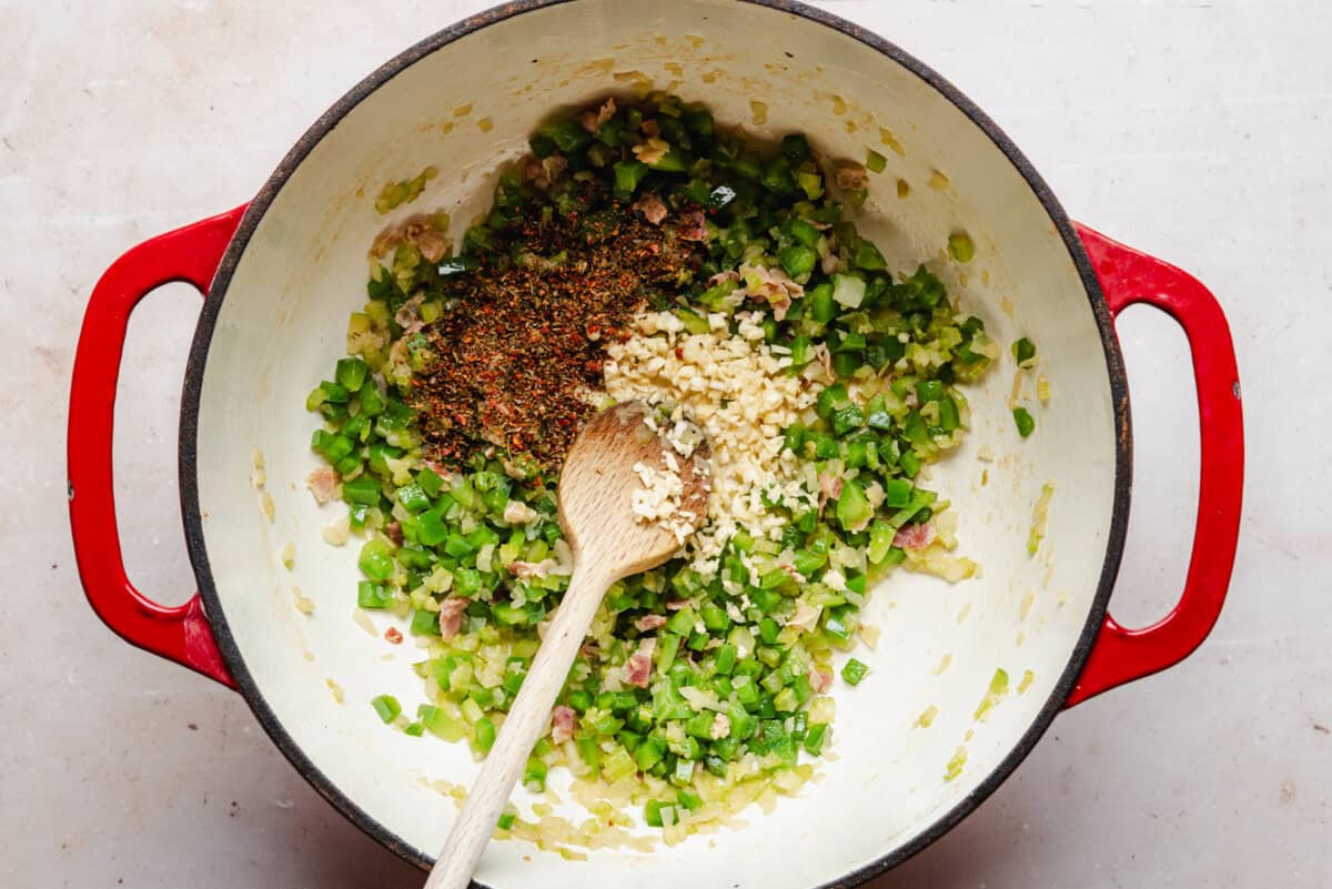 A red Dutch oven contains sautéed diced green bell peppers, onions, and bacon, with minced garlic and dried spices on top. A wooden spoon rests inside the pot, ready for stirring.