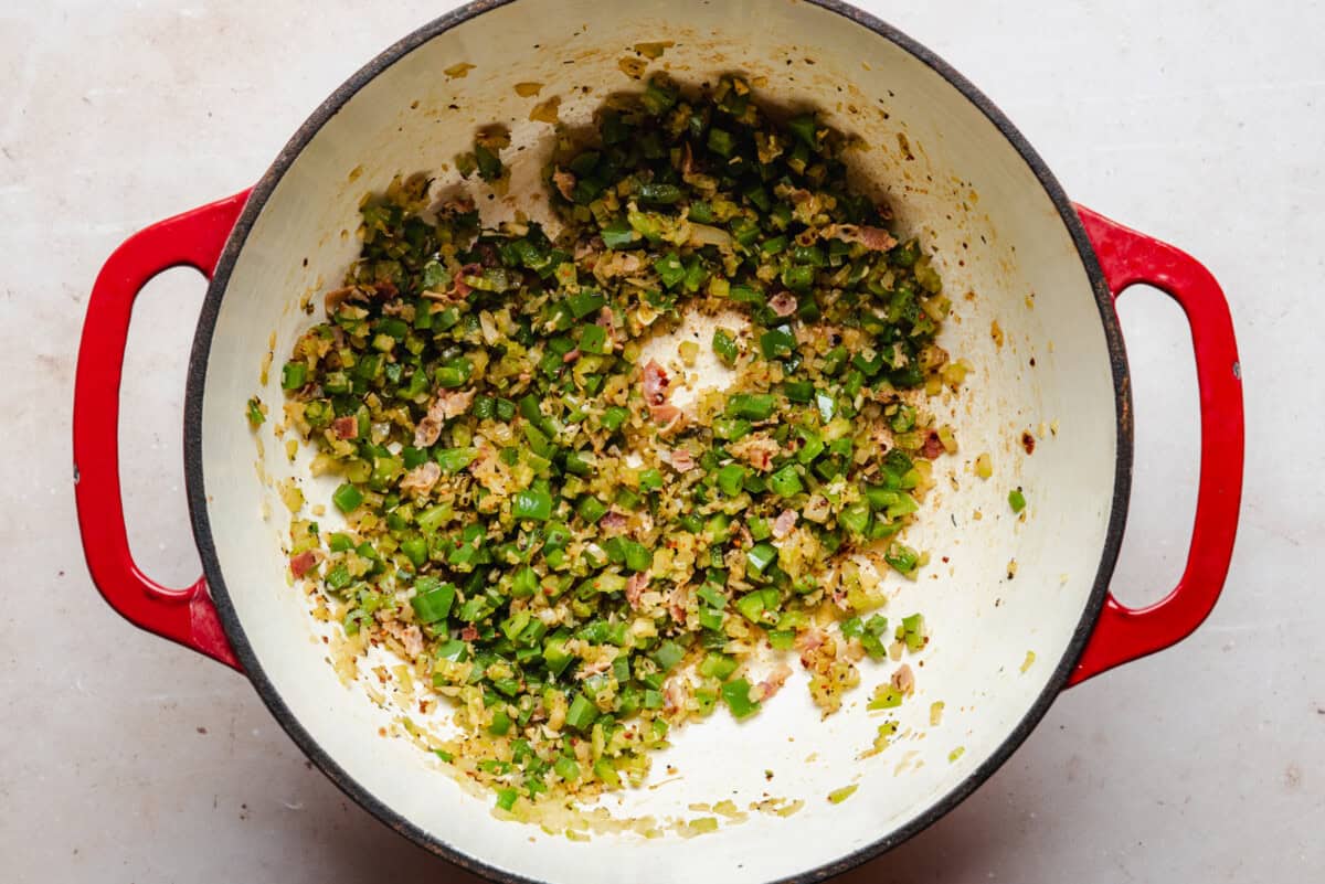 Chopped green bell peppers and onions sautéing with minced meat in a red-handled white Dutch oven on a light-colored surface.