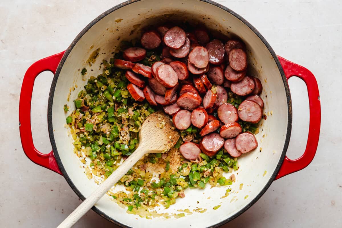 A red Dutch oven contains sliced sausage, chopped green bell peppers, onions, and seasonings being sautéed. A wooden spoon rests inside the pot, mixing the ingredients.