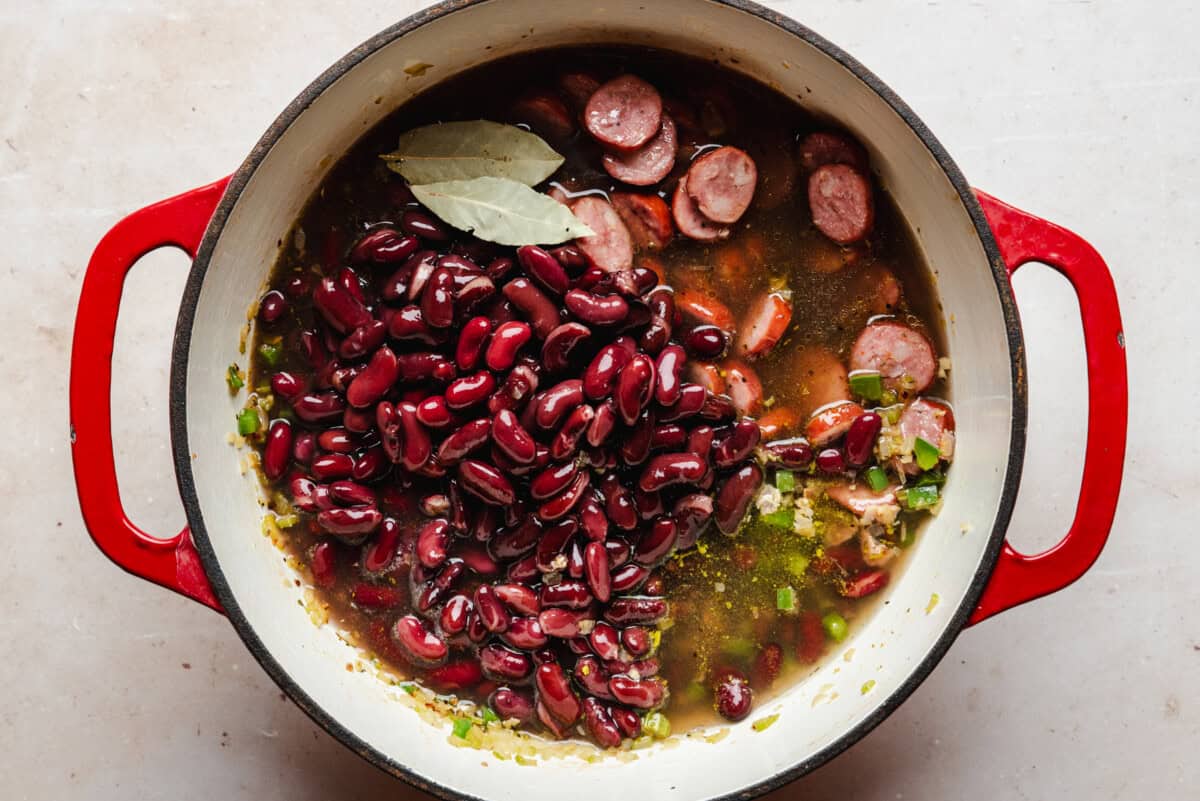 A red pot filled with sliced sausage, kidney beans, broth, chopped vegetables, and two bay leaves, ready to be cooked on a light-colored countertop.