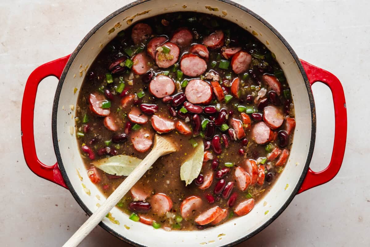 A red pot filled with a stew of sliced sausage, kidney beans, chopped green vegetables, and two bay leaves, with a wooden spoon resting inside.