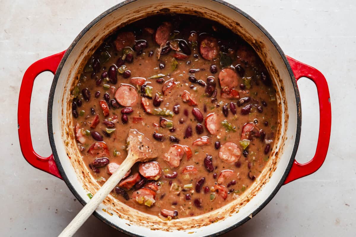 A red Dutch oven filled with a stew of sliced sausage, red beans, chopped vegetables, and a thick brown broth, with a wooden spoon resting inside.