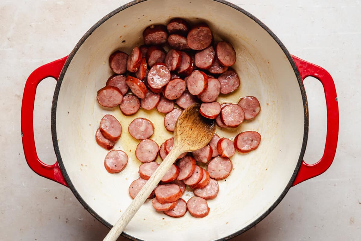A red-handled pot on a light surface contains sliced sausage being cooked, with a wooden spoon resting inside. The sausage slices are browning in oil.