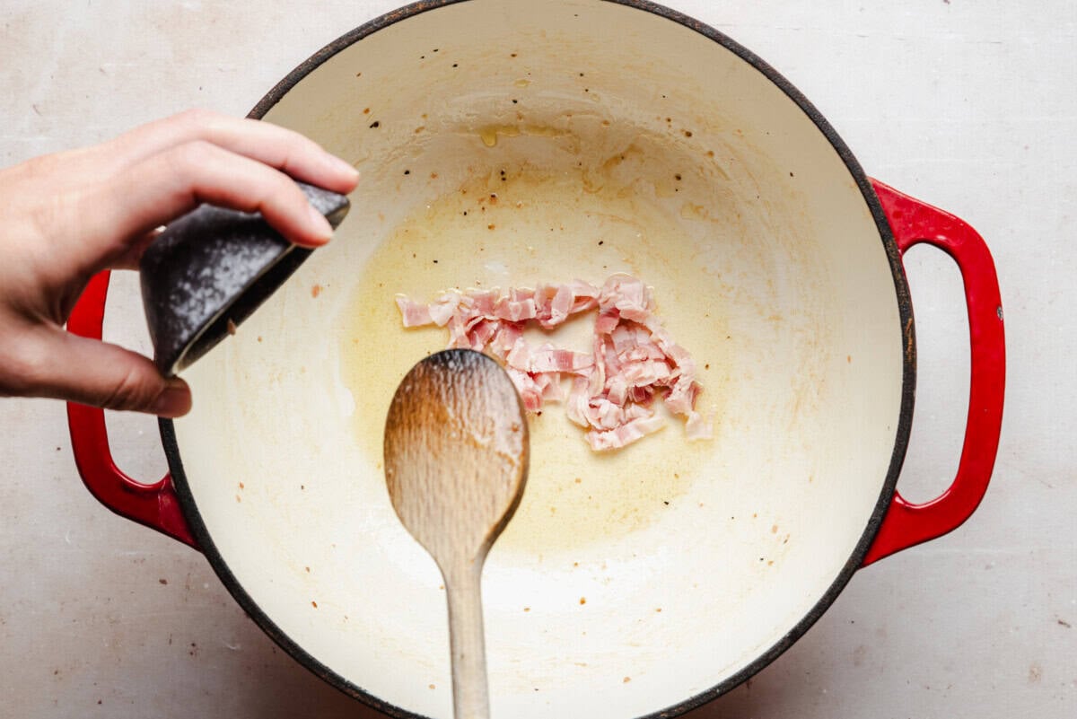 A hand sprinkles salt into a red Dutch oven with chopped bacon pieces cooking in oil, while a wooden spoon stirs the ingredients.