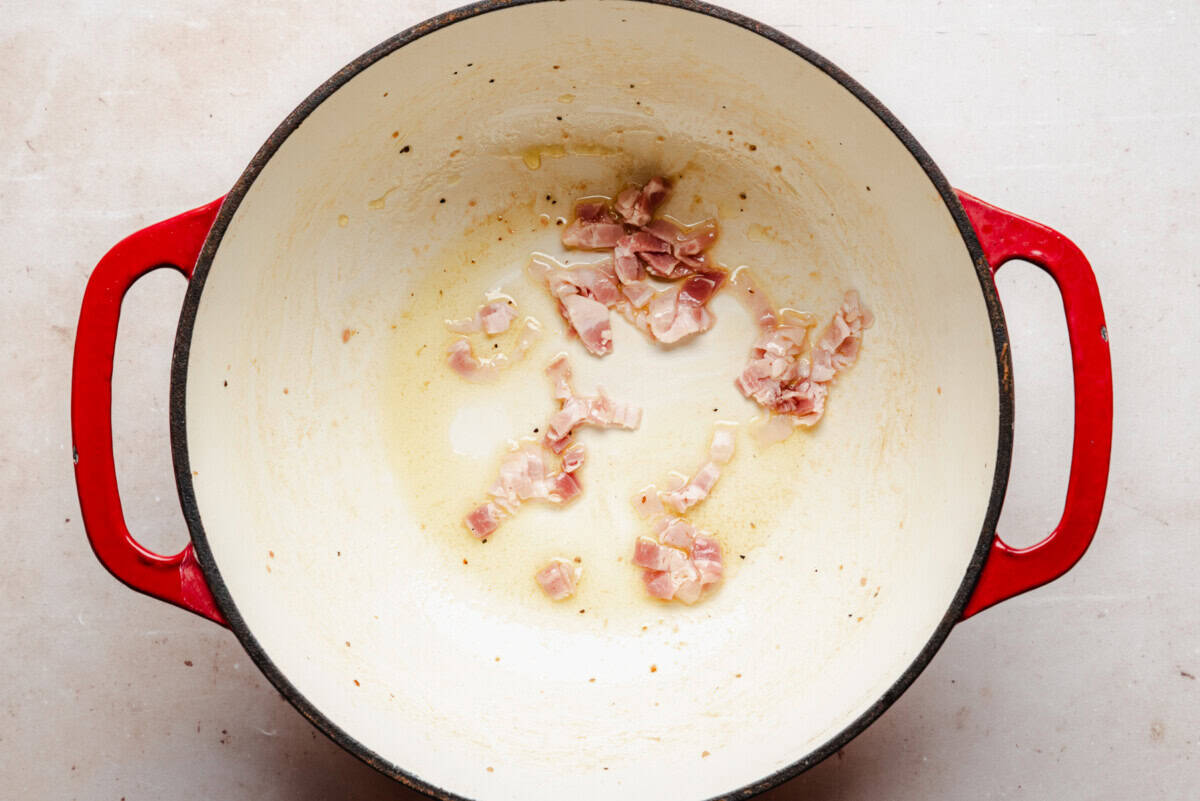 A few pieces of chopped bacon are sautéing in a large, white enameled pot with red handles, set on a light-colored surface.