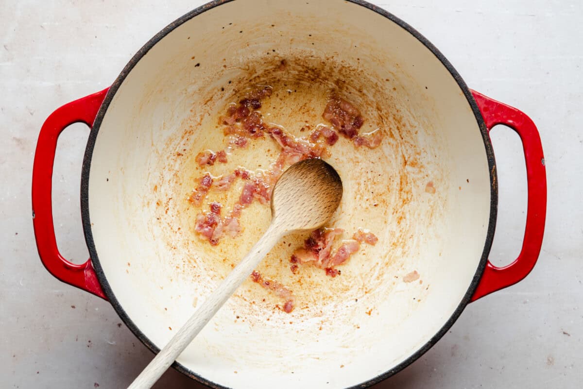 A red-handled pot contains partially cooked bacon pieces being stirred with a wooden spoon. The pot shows some browning and rendered fat, sitting on a light-colored surface.