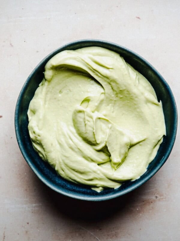 A small dark bowl filled with smooth, creamy light green avocado crema sits on a light-colored surface.